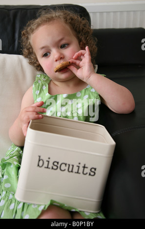 Une fillette de 3 ans assis sur le canapé à la maison manger des cookies à partir d'un excès d'étain et de biscuit Banque D'Images