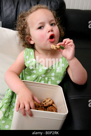 Une fillette de 3 ans assis sur le canapé à la maison étant greeding et eatings beaucoup de cookies et biscuits Banque D'Images