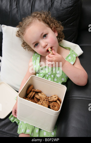 Une fillette de 3 ans assis sur le canapé à la maison étant greeding et eatings beaucoup de cookies et biscuits Banque D'Images