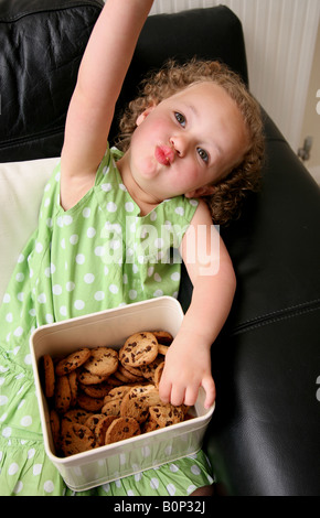 Un vilain 3 ans, fille, assise sur le canapé à la maison manger des biscuits Banque D'Images