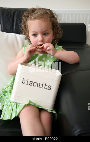 Une fillette de 3 ans assis sur le canapé à la maison manger des cookies à partir d'un excès d'étain et de biscuit Banque D'Images