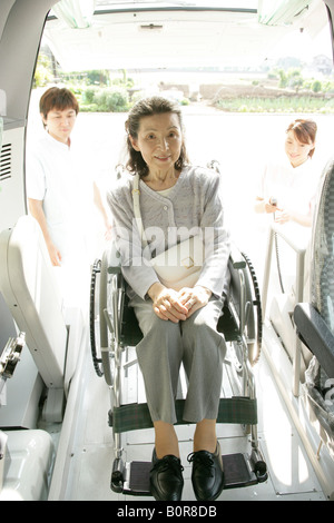 Senior woman sur fauteuil roulant d'entrer dans voiture, deux infirmières en arrière-plan Banque D'Images