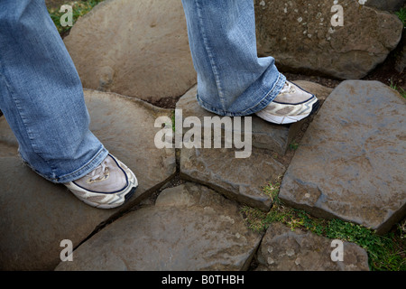 Jambes de femme portant des jeans et des vieilles baskets sur basalte rouge rock formations hexagonales à la Chaussée des géants du nord du comté d'Antrim Banque D'Images