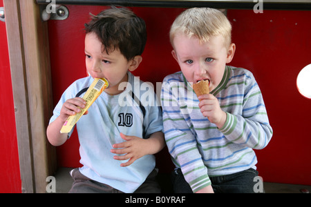 Deux 3 ans garçons assis dans une escalade dans un parc un manger un lolly et l'un manger une glace Banque D'Images