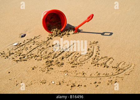 Vue horizontale d'un seau en plastique rouge et la cosse sur la plage à côté de 'vacances' écrit dans le sable Banque D'Images