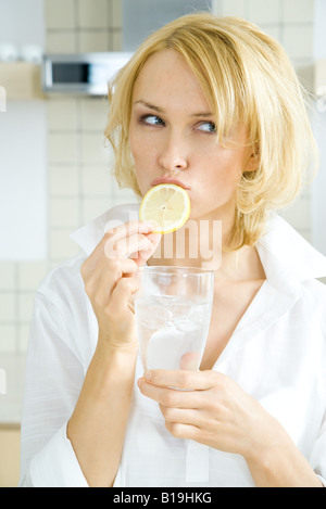 Jeune femme mordre dans une tranche de citron, à l'écart, tenant un verre d'eau Banque D'Images