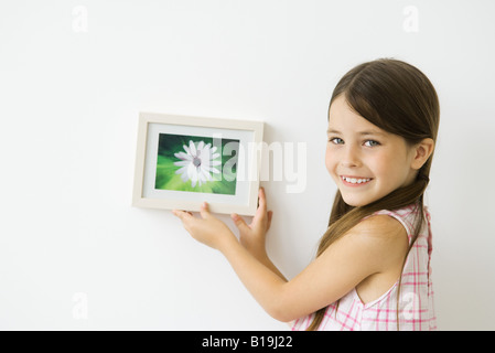 Little girl holding photo encadrée contre mur, smiling over Shoulder at camera Banque D'Images