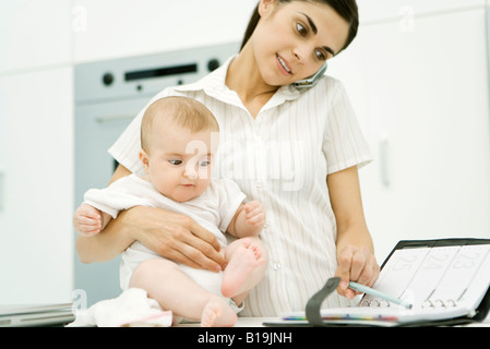 Woman holding baby, à l'aide d'un téléphone cellulaire et à la recherche à l'ordre du jour Banque D'Images