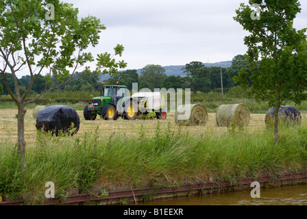 Tracteur John Deere vert tirant une ramasseuse-presse à balles rondes de foin alors que dans un champ sur une ferme de Cheshire Banque D'Images