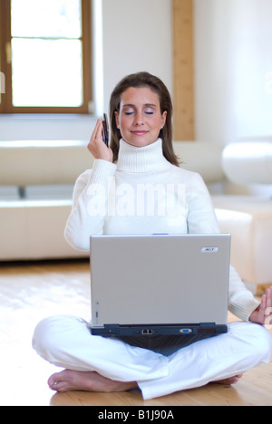 Young attractive woman practicing yoga avec un ordinateur portable sur ses genoux et le mobile dans la main, assis sur le plancher dans le salon Banque D'Images