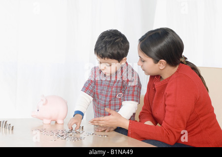 Boy making des piles de pièces de monnaie près de une tirelire avec sa mère assis à côté de lui Banque D'Images