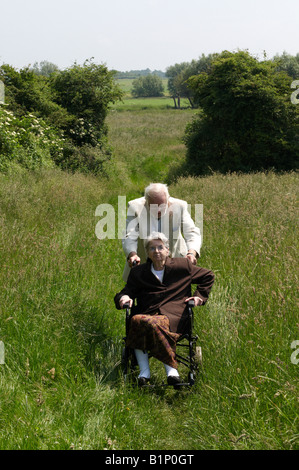 Vieille dame en fauteuil roulant dans la campagne avec son mari âgé essayant de pousser sa place une banque herbeux Cambridge Banque D'Images