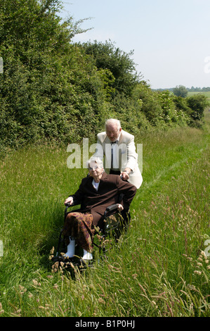 Vieille dame en fauteuil roulant dans la campagne avec son mari âgé essayant de pousser sa place une banque herbeux Cambridge Banque D'Images