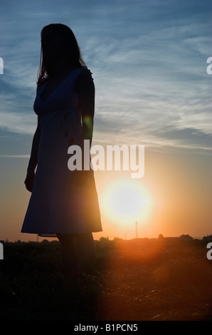 Silhouette of a young woman standing in field robe d'été à la tombée du soleil derrière Banque D'Images