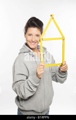 Woman face à la caméra, tenant dans ses mains un mètre pliant jaune repliée en forme de maison Banque D'Images