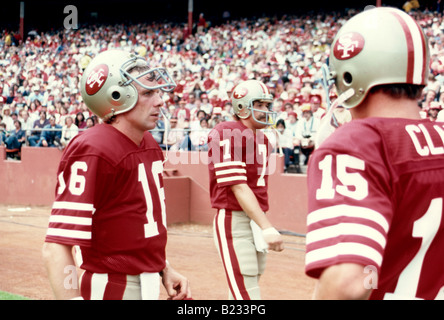 Joe Montana porte un maillot rouge et un casque doré sur la ligne de touche aux côtés de ses coéquipiers Guy Benjamin et Dwight Clark dans un stade bondé. Banque D'Images