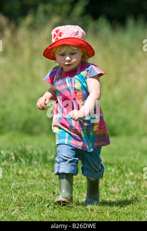 Une jeune fille cute toddler running walking portant des bottes et un chapeau de soleil rouge sur une journée ensoleillée dans un champ vert. Banque D'Images
