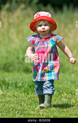 Une jeune fille cute toddler running walking portant des bottes et un chapeau de soleil rouge sur une journée ensoleillée dans un champ vert. Banque D'Images