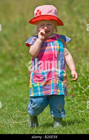 Une jeune fille cute toddler pleurer portant wellies et un soleil rouge hat lors d'une journée ensoleillée dans un champ vert. Banque D'Images