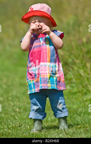 Une jeune fille cute toddler pleurer portant wellies et un soleil rouge hat lors d'une journée ensoleillée dans un champ vert. Banque D'Images