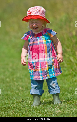 Une jeune fille cute toddler pleurer portant wellies et un soleil rouge hat lors d'une journée ensoleillée dans un champ vert. Banque D'Images