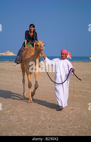 Jeune femme d'origine indienne omanais rides un chameau mené par l'homme sur l'Oman Al Sawadi Beach Banque D'Images