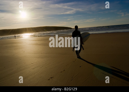 Un surfeur surf avec promenades sous les bras de la mer au coucher du soleil tourné sur une plage à Cornwall en Angleterre avec un ciel magnifique Banque D'Images