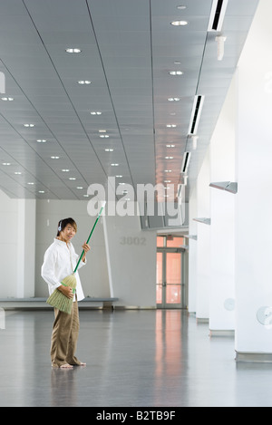 Homme debout dans le hall, playing air guitar avec balai, listening to headphones, smiling at camera Banque D'Images