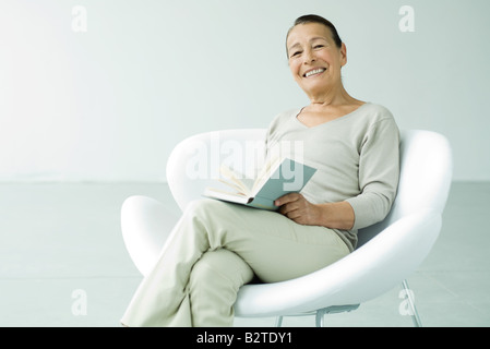 Senior woman sitting in chair, holding book, smiling at camera Banque D'Images