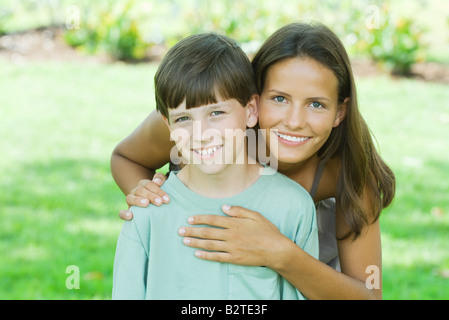 Teen girl debout derrière son jeune frère, les mains sur l'épaule et le coeur, les deux smiling at camera