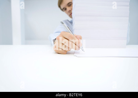 Homme assis derrière grande pile de documents, écrit sur une seule feuille de papier Banque D'Images