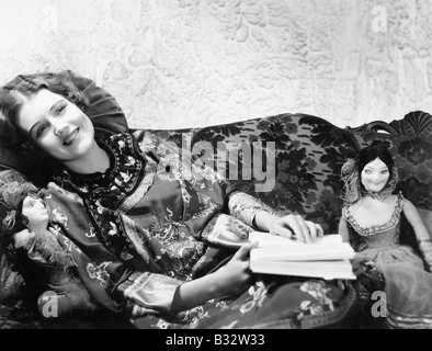 Femme dans une robe chinoise allongée sur le canapé avec ses poupées et un livre Banque D'Images