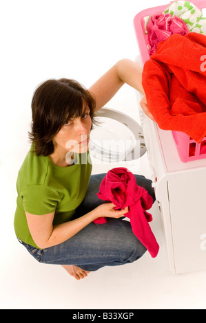 Jeune femme caucasienne portant un t-shirt vert et un Jean chargeant un lave-linge domestique avec des vêtements de couleurs vives provenant d'un panier à linge. Banque D'Images
