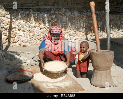 Le Kenya, l'Ile de Pate, Faza. Une femme Swahili grain winnows devant sa maison tandis que son jeune fils tient un grand mortier et pilon à côté d'elle. Sa maison, comme la plupart des autres dans le village, est fait de chiffon corail.L'histoire mouvementée de Faza date de plusieurs centaines d'années. Il a été détruit par Pate au 13ème siècle et encore une fois par les Portugais en 1586. Les habitants puis leur allégeance aux Portugais au 17ème siècle dans une tentative de soumettre la pate gênants. Faza a diminué en importance dans les 18e et 19e siècles et est maintenant un bras mort. Banque D'Images