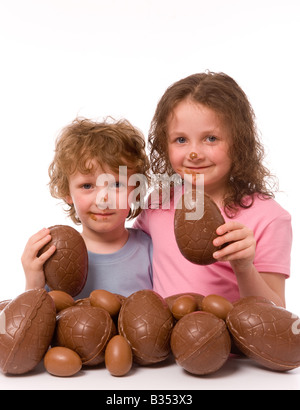 2 jeunes enfants avec du chocolat sur leur visage, chacun tenant un morceau d'oeuf de chocolat, avec beaucoup plus d'oeufs de Pâques au premier plan. Arrière-plan blanc Banque D'Images