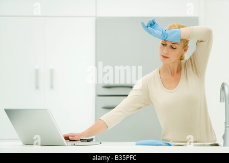 Femme debout à l'évier de cuisine, atteignant pour ordinateur portable, portant des gants de caoutchouc Banque D'Images