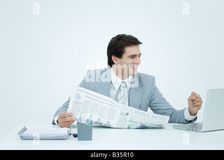 Young businessman looking at laptop, holding newspaper et clenching fist Banque D'Images