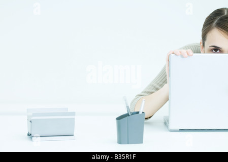 Jeune femme peeking over laptop, portrait Banque D'Images