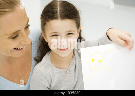 Girl holding up et montrant le dessin du soleil, mère souriant derrière sa fille Banque D'Images