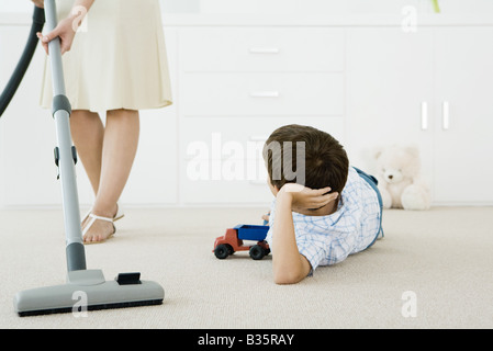 Garçon allongé sur le sol avec des jouets, regardant sa mère l'aspirateur autour de lui Banque D'Images