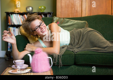 Young woman lying on sofa at home eating cookies et boire le thé Banque D'Images