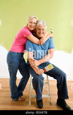 Portrait of smiling couple assis en face de la moitié mur peint avec les pinceaux Banque D'Images