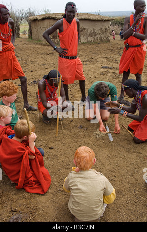 Kenya, Masai Mara National Reserve. Au cours d'une visite d'une manyatta Masaï enfants sur une famille safari regardez comme les guerriers démontrer faire feu en frottant des bâtons. Banque D'Images