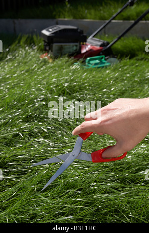 Mans part fin de l'adolescence dans la jeune vingtaine, couper l'herbe dans un jardin avec une paire de ciseaux avec tondeuse à essence à l'arrière-plan Banque D'Images