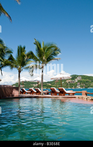 La Casa que Canta Resort Pool Zihuatanejo Mexique | ZIHUATANEJO, Mexique — des palmiers encadrent une piscine à débordement à la Casa que Canta, un complexe situé à flanc de falaise au-dessus de la baie de Zihuatanejo sur la côte Pacifique du Mexique. Des rangées de chaises longues ombragées de parasols bordent la terrasse de la piscine, avec les collines boisées et le développement du front de mer de Zihuatanejo visibles de l'autre côté de la baie. Banque D'Images
