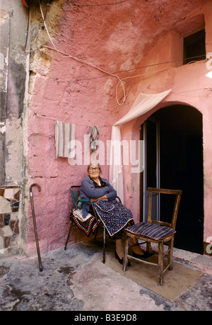 Une vieille femme EN TABLIER EST ASSIS SUR UNE CHAISE AVEC UNE CANNE APPUYÉE CONTRE LE MUR D'UNE ARCHE ROSE Banque D'Images