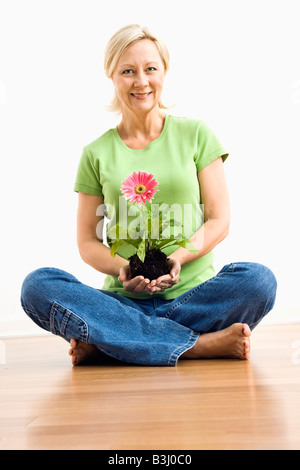 Portrait of smiling hot blonde woman sitting on floor holding gerber daisy plant rose Banque D'Images