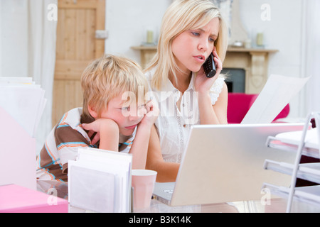 Femme à l'aide de téléphone en home office avec ordinateur portable tout en jeune garçon attend Banque D'Images