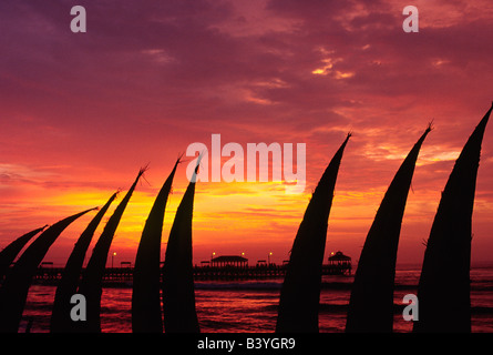 Le Pérou, La Libertad, le soleil peint le ciel rouge, silhouetting totora (Reed) bateaux empilés sur le front de mer à Huanchaco, Banque D'Images