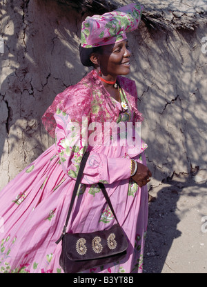 La Namibie, Damaraland, Palmwag. Une Femme Herero se tient dans l'ombre à l'extérieur de sa maison. Les origines de sa robe élaborée et unique style hat remonte au 19ème siècle les missionnaires allemands qui se sont opposés à ce qu'ils considéraient comme une forme de modestie parmi la tribu de robe. Ils ont insisté pour que les femmes Herero peu pratique d'adopter leur propre style de robes de l'époque victorienne. Au fil des ans, robes Herero colorés ont évolué aussi énorme crinolines porté sur cinq jupons. Banque D'Images